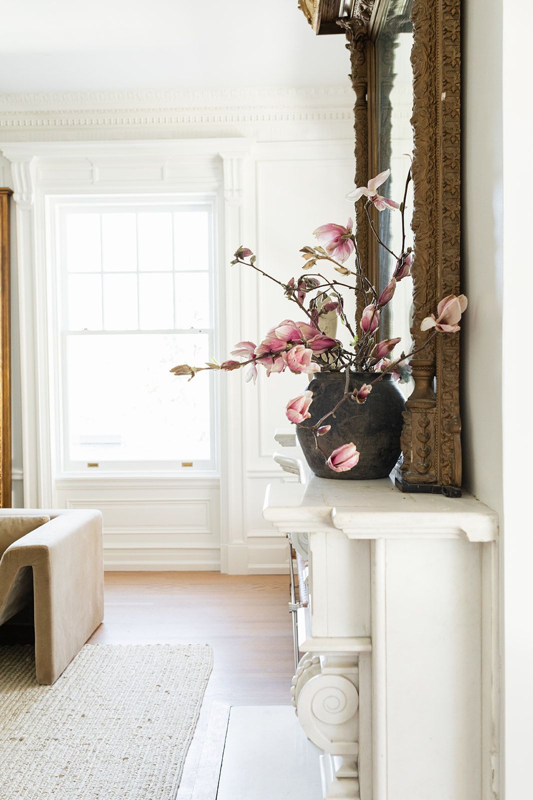 Pink cherry blossoms in black vase on white mantel near bright window.