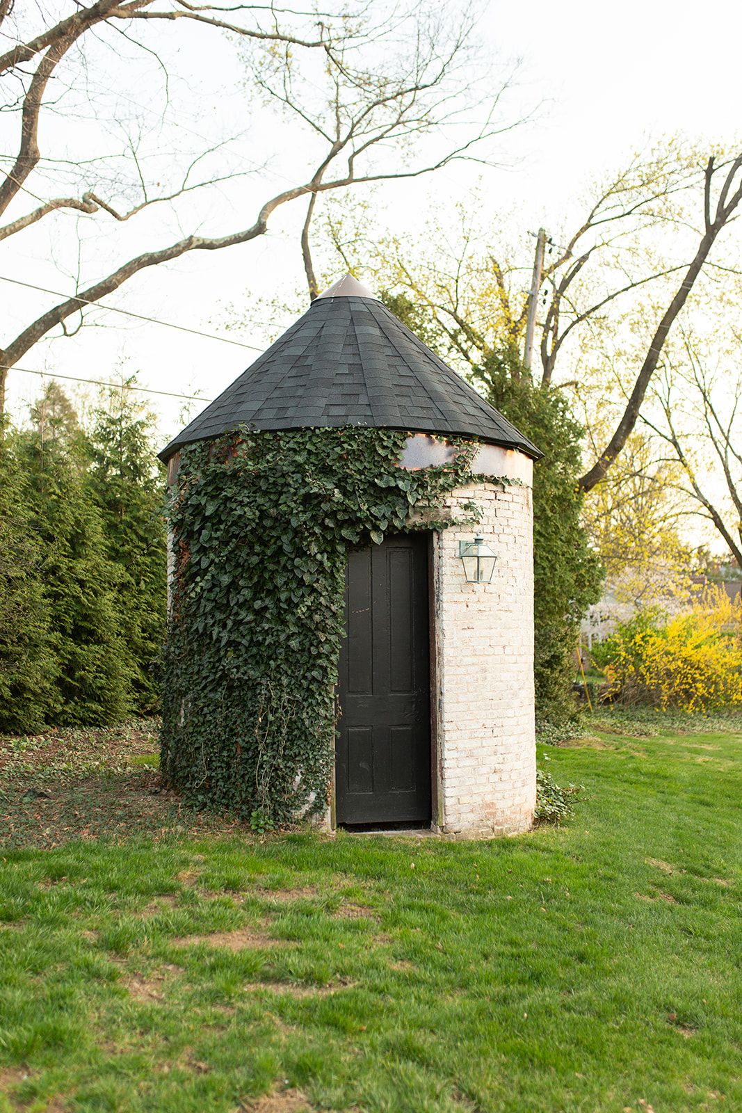 Small white circular garden shed with black door and climbing ivy on exterior sits on grassy lawn.