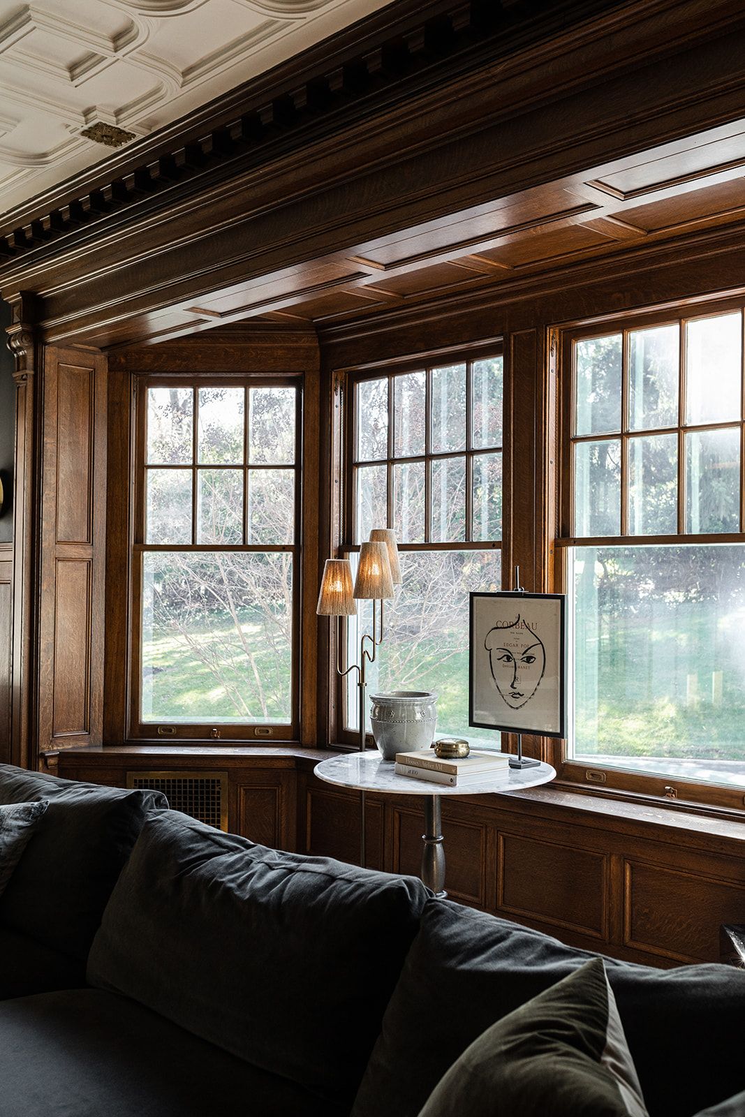 Cozy window nook with dark wood paneling, large windows, and gray sofa overlooking garden.