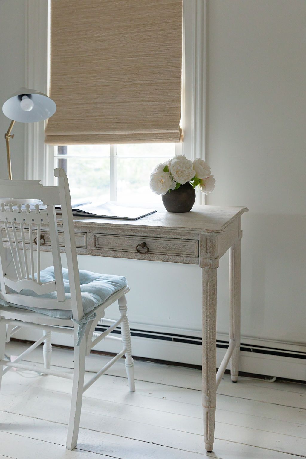 Elegant white vintage desk and chair against a window with natural roller shade and white peonies in a vase.