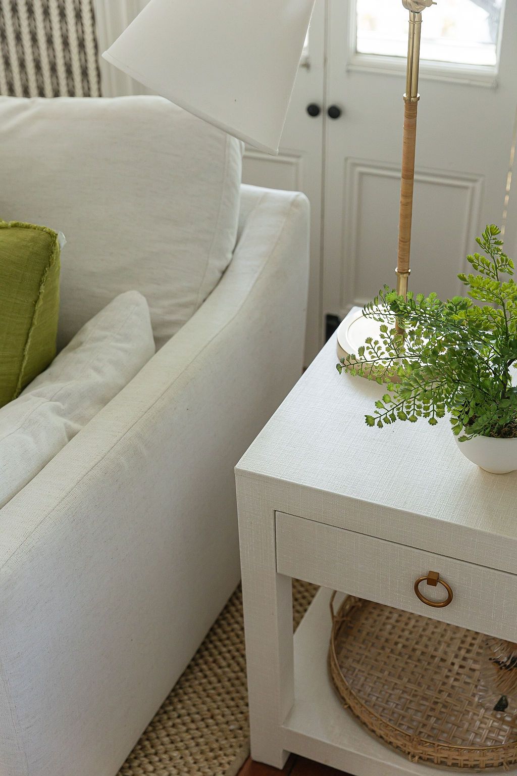 White living room furniture with green accent pillows and a potted plant on small white side table near a doorway.