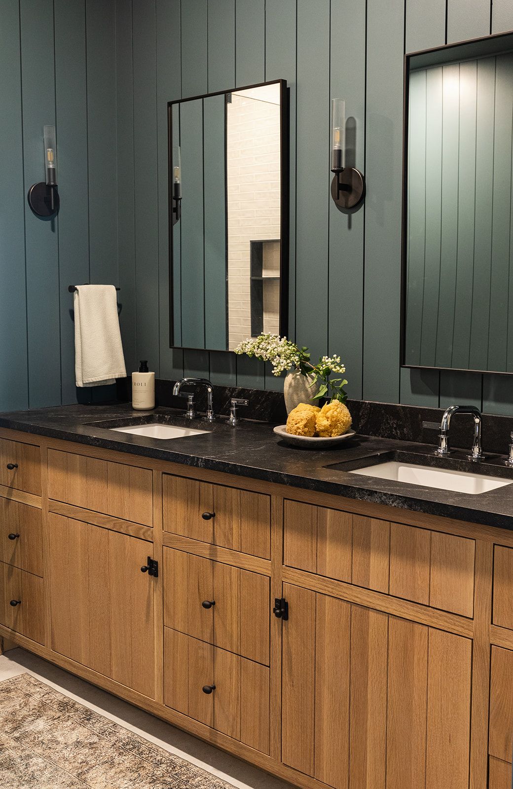 Modern bathroom vanity with wooden cabinets, black countertop, dual sinks and sage green shiplap walls with sconces.