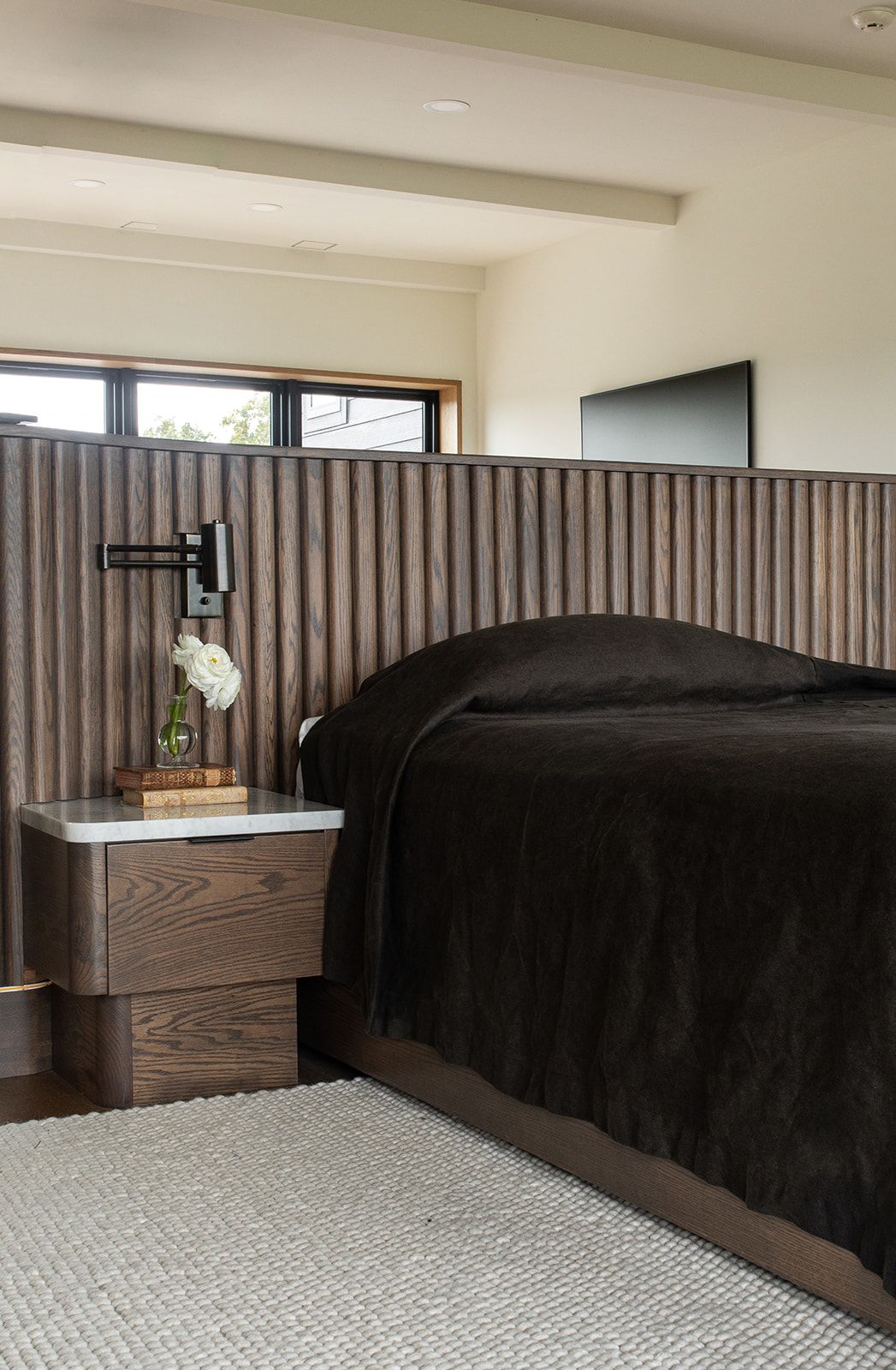 Modern bedroom with tall fluted wood headboard, dark bedding, wooden nightstand, and wall-mounted lamp in neutral tones.