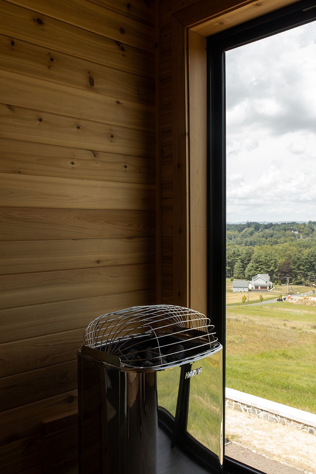 A modern sauna interior with wood-paneled walls, a sleek electric heater by a large window overlooking a green rural landscape.