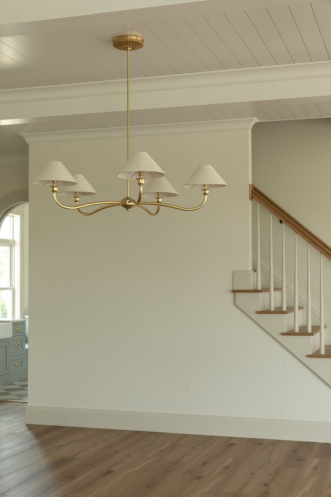 Elegant brass chandelier with white shades in an open foyer featuring hardwood floors and a classic staircase.