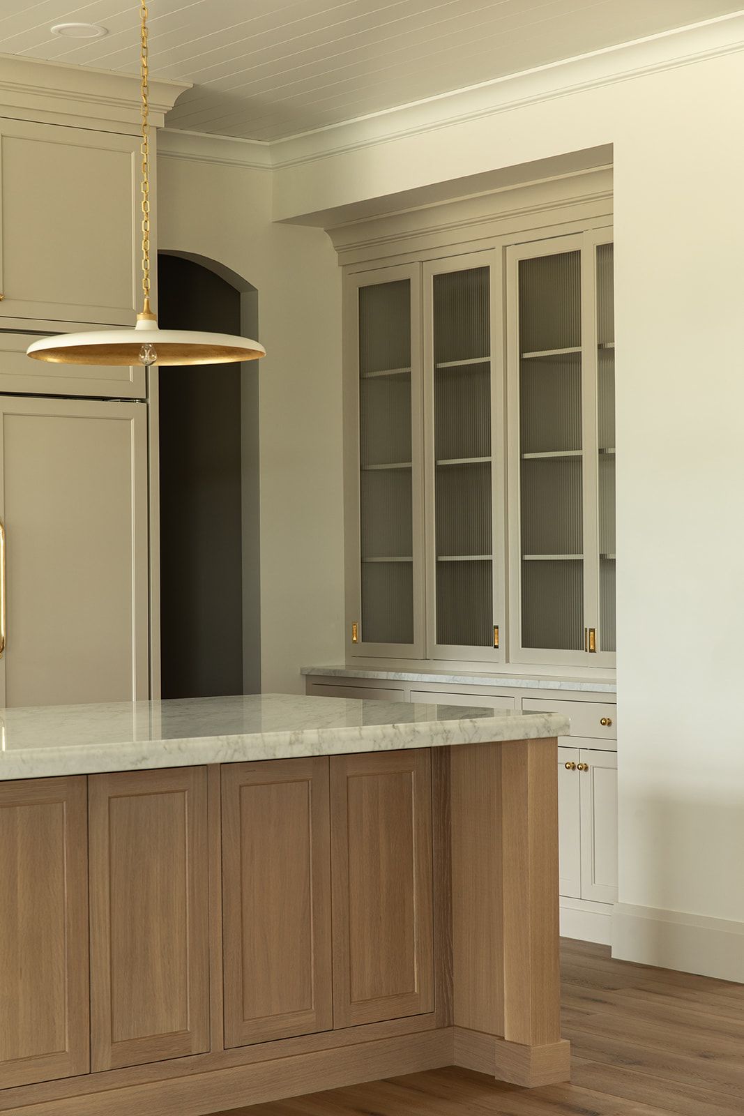 Transitional kitchen with greige cabinets, glass-front upper cabinets, brass hardware, natural wood island, and marble countertops.