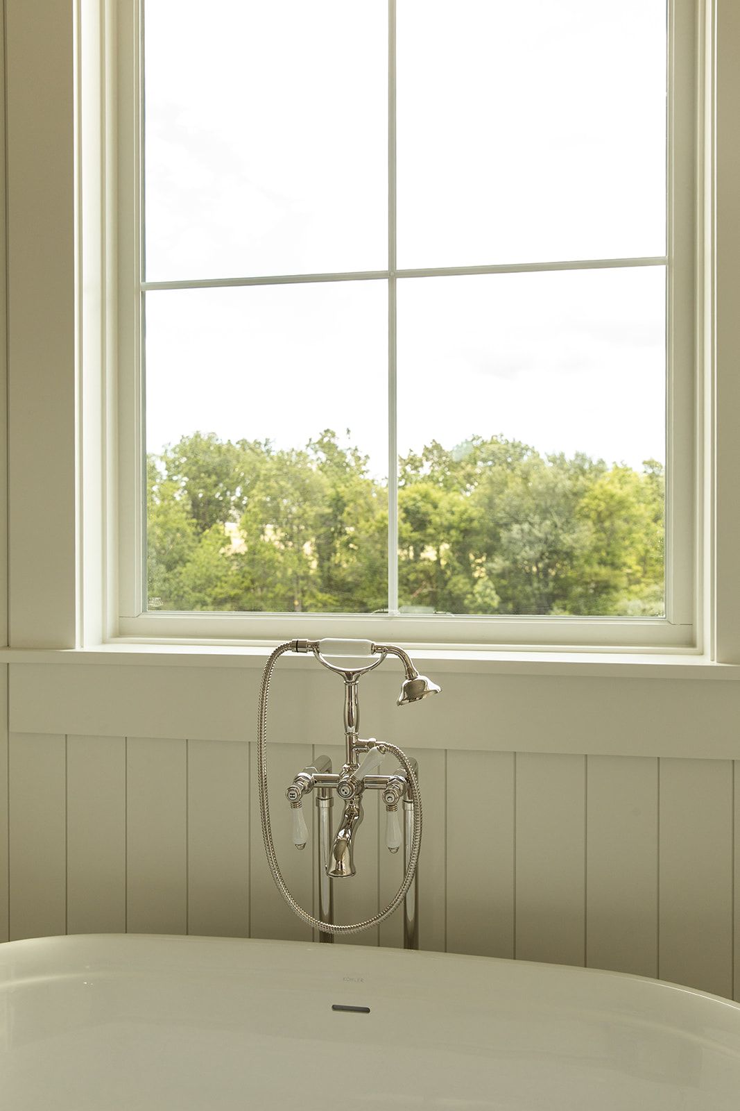 Freestanding soaking tub with chrome floor-mount faucet beside a large window overlooking lush green trees.