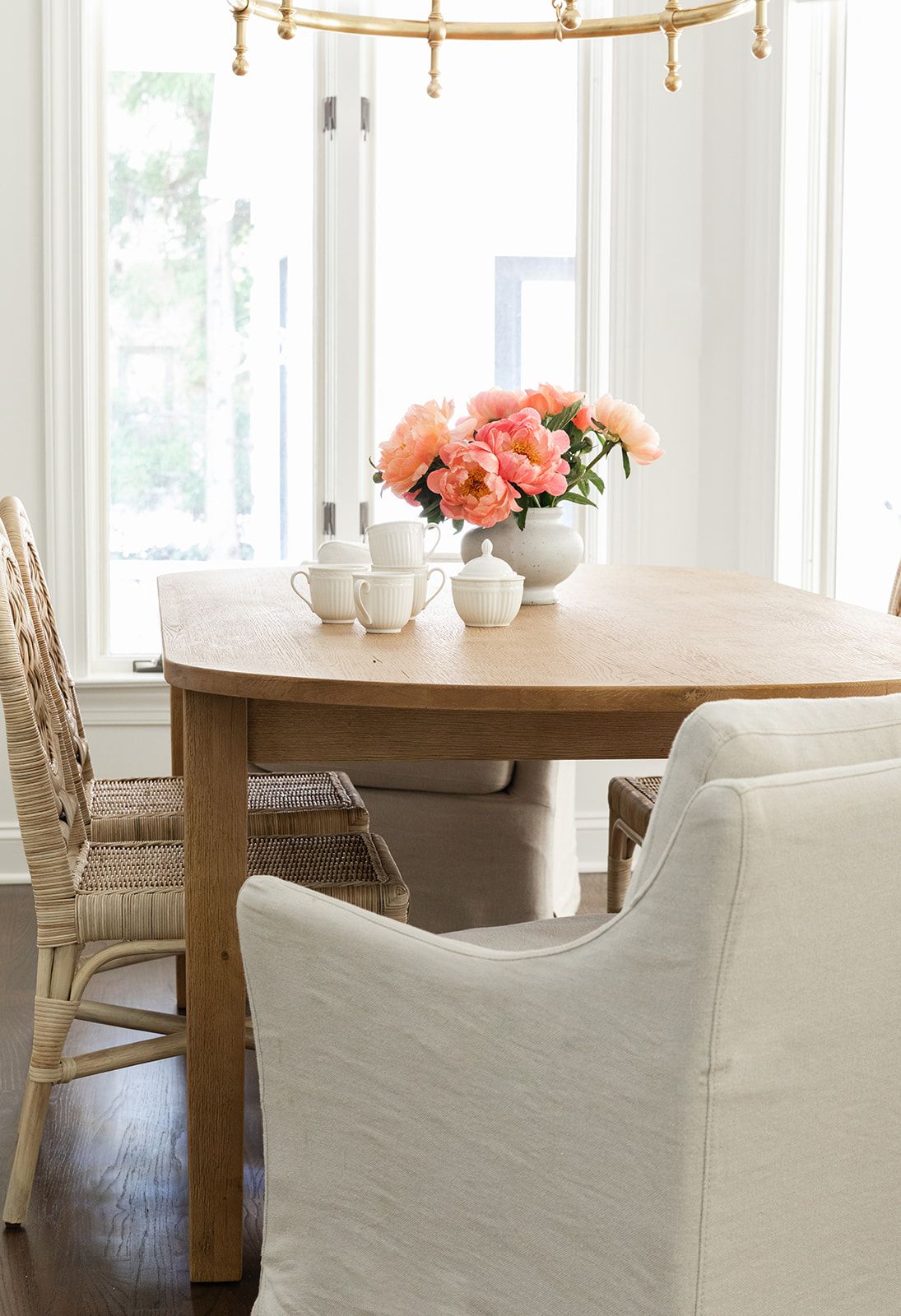 Bright dining room with oval wood table, white slipcovered chairs, rattan seats, and pink peonies in a white vase.