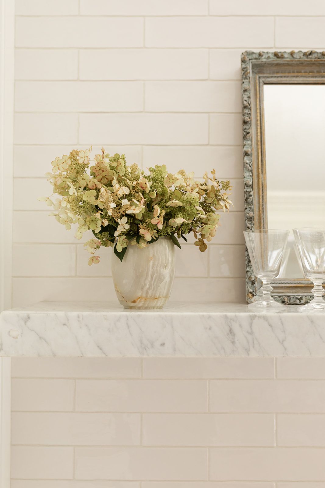 Cream hydrangeas in marble vase on white shelf with vintage silver frame mirror.