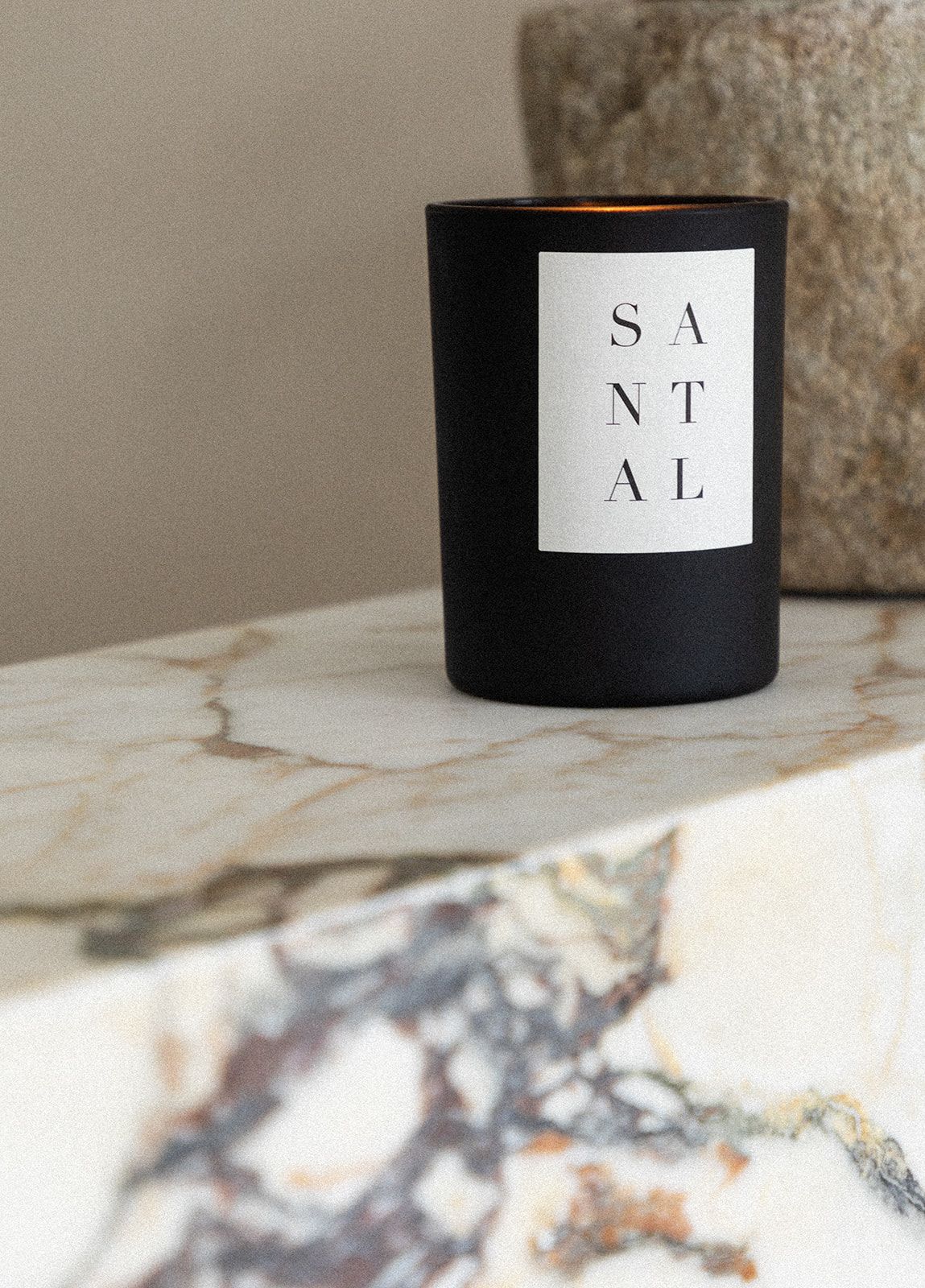 Minimalist black candle with 'SANTAL' label sits on marble surface in soft natural lighting.