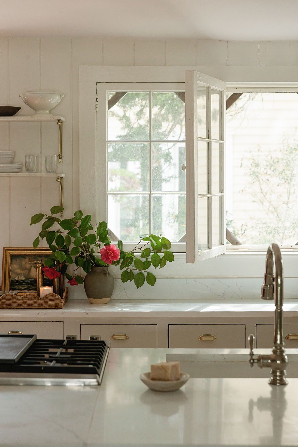 Modern white kitchen with bright windows, green plants, and clean countertops featuring vintage-style brushed metal fixtures.