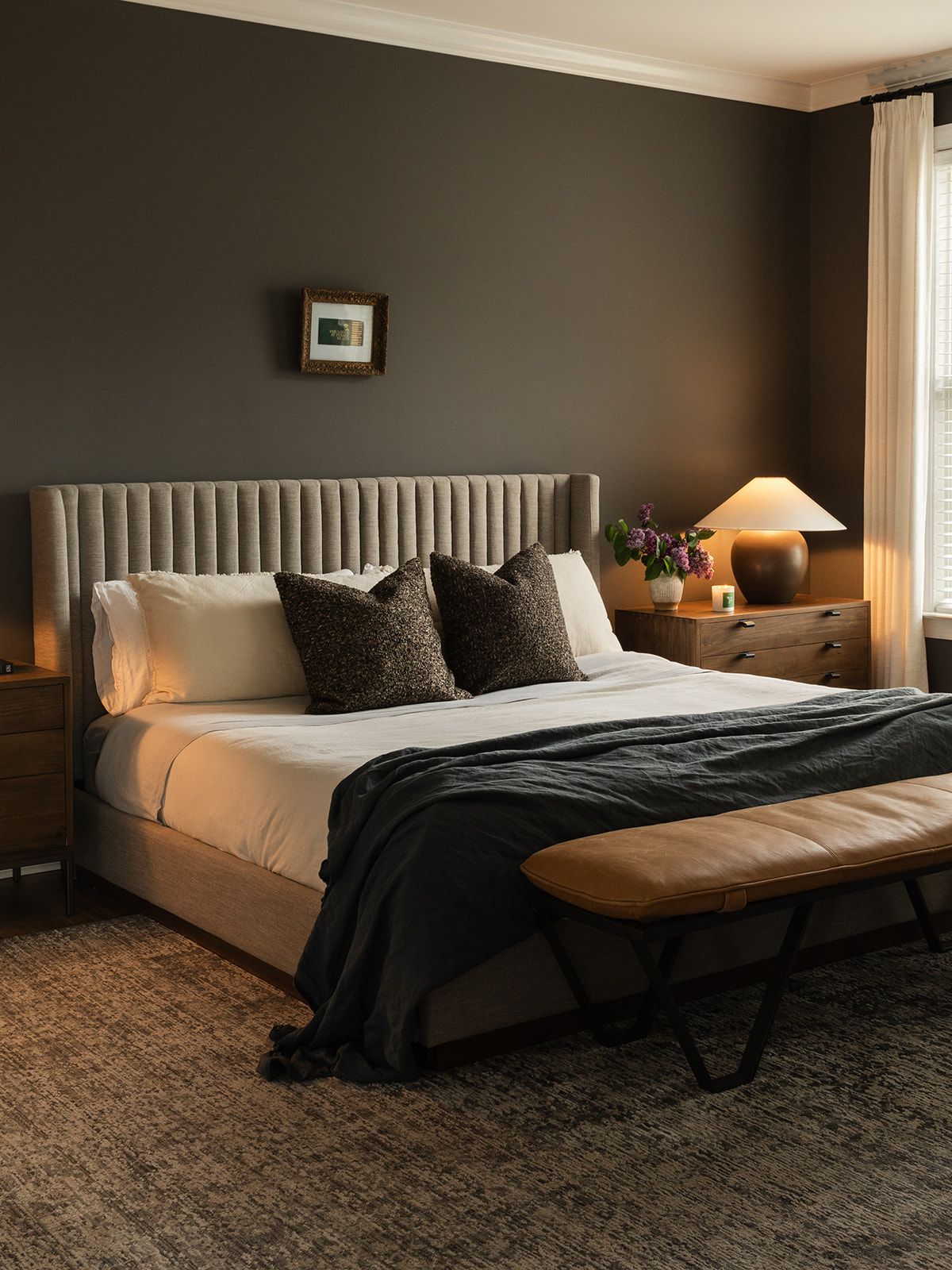 Modern bedroom with dark walls featuring an upholstered bed with white linens, black throw pillows, and warm ambient lighting.
