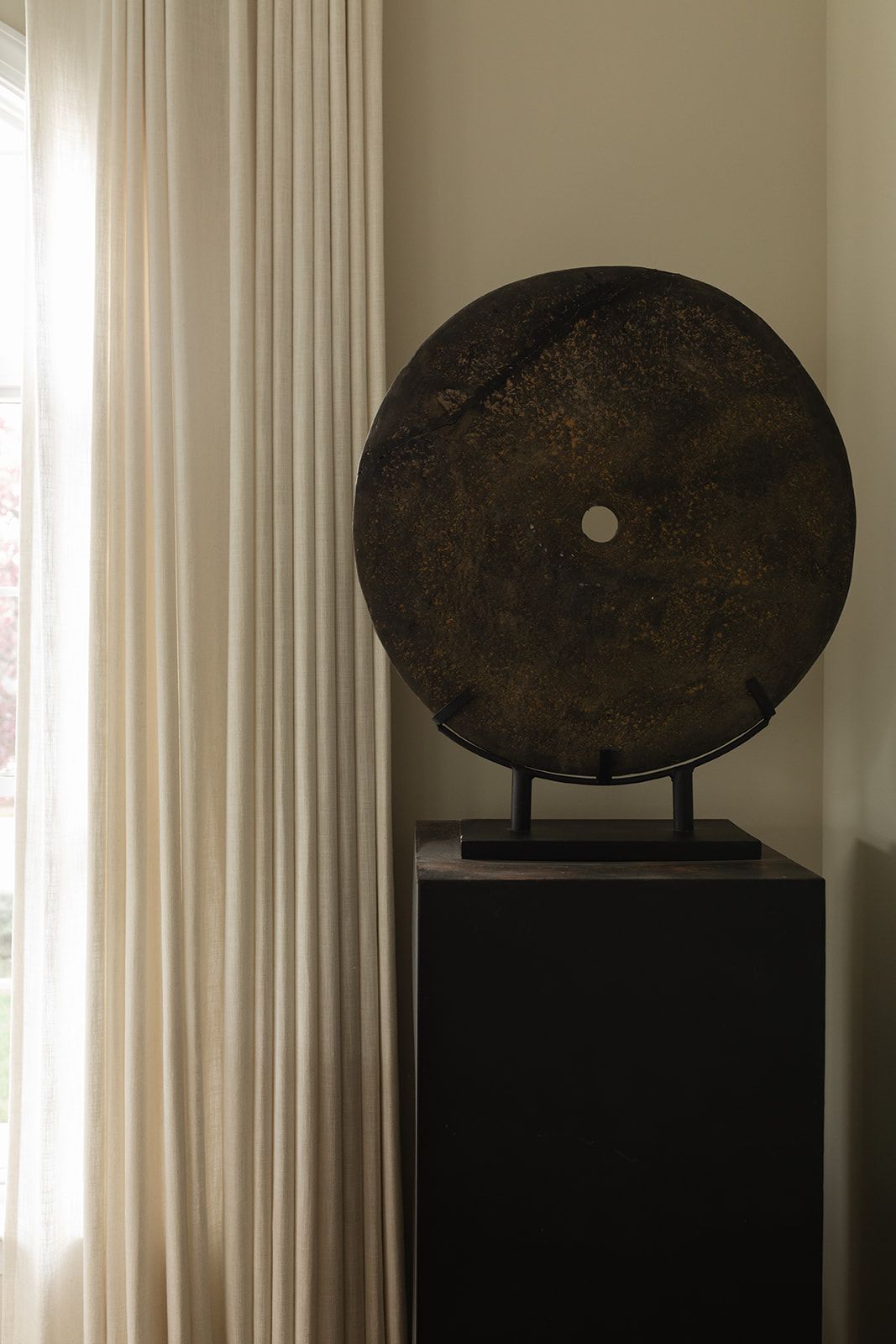Abstract oval sculpture displayed on black pedestal against light curtained window.