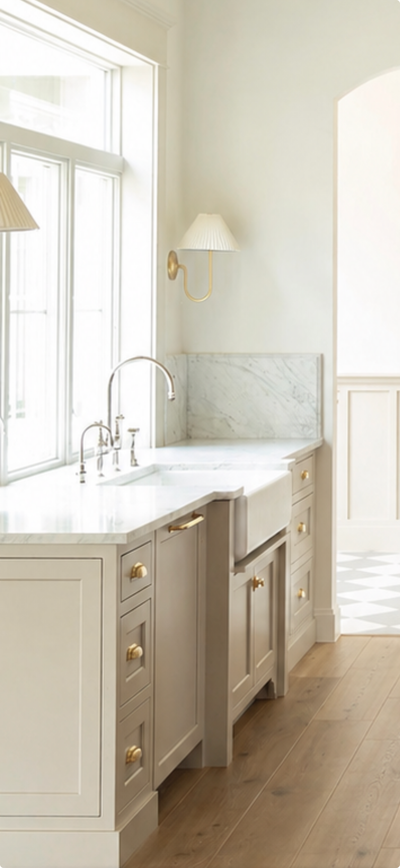 Elegant kitchen with taupe shaker cabinets, brass hardware, marble backsplash, farmhouse sink, and gold wall sconce on white walls.