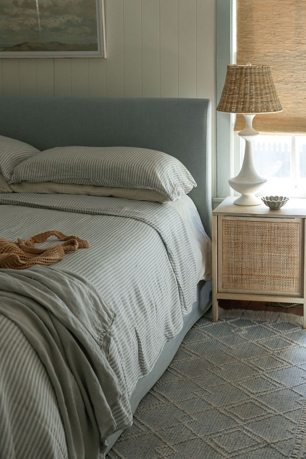 A cozy bedroom with gray bedding, woven nightstand, white table lamp, and geometric patterned area rug.