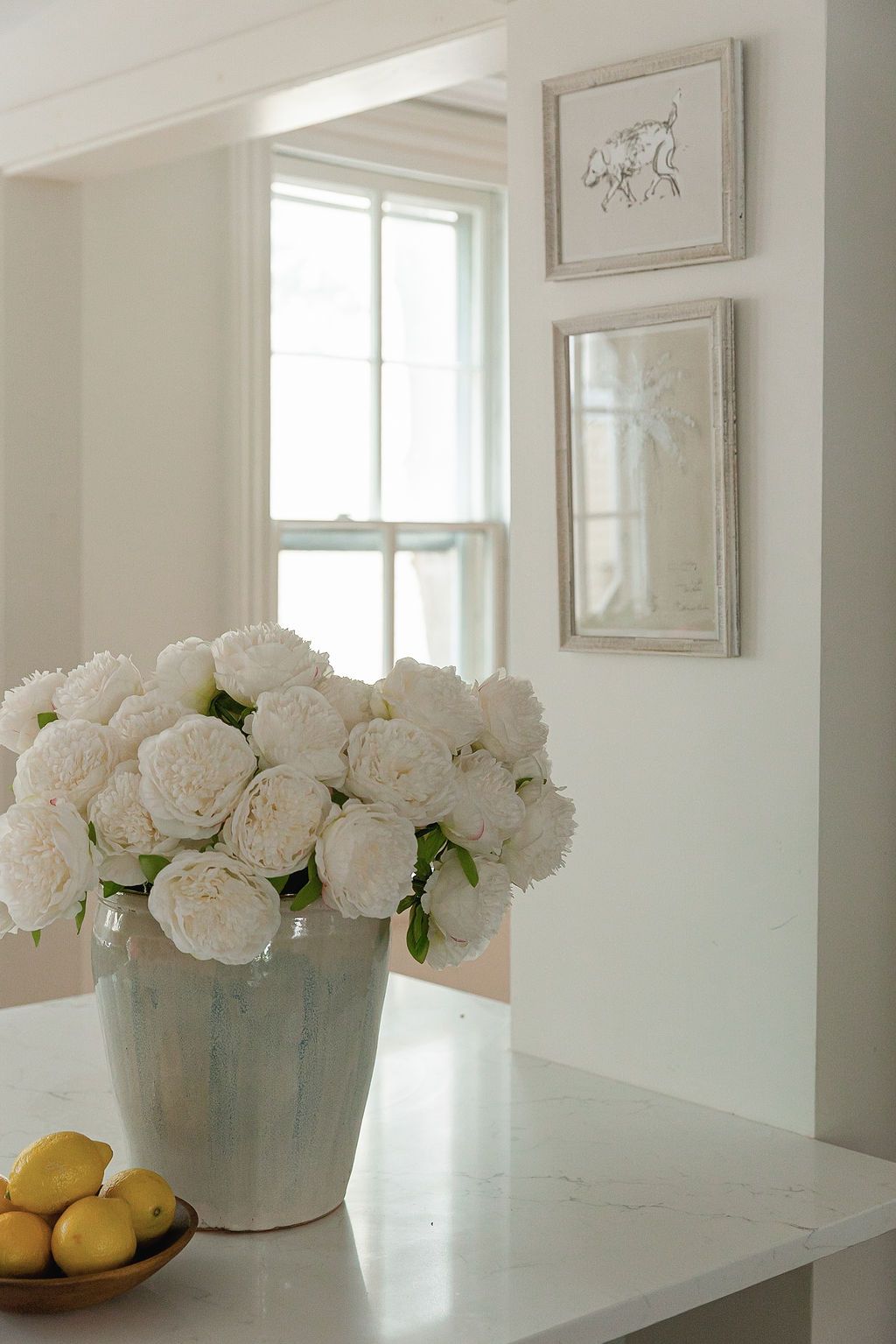 Large white peony arrangement with lemons on marble counter below framed artwork.