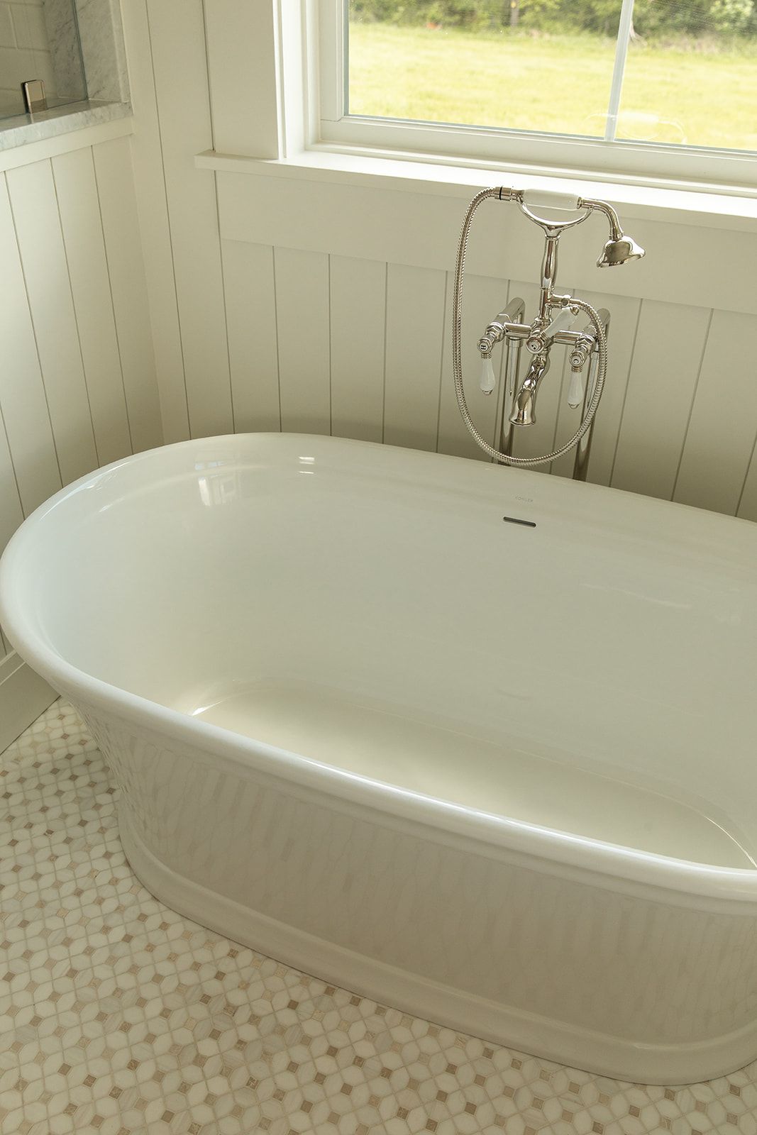Freestanding white soaking bathtub with chrome floor-mount faucet beside a bright window with garden views.