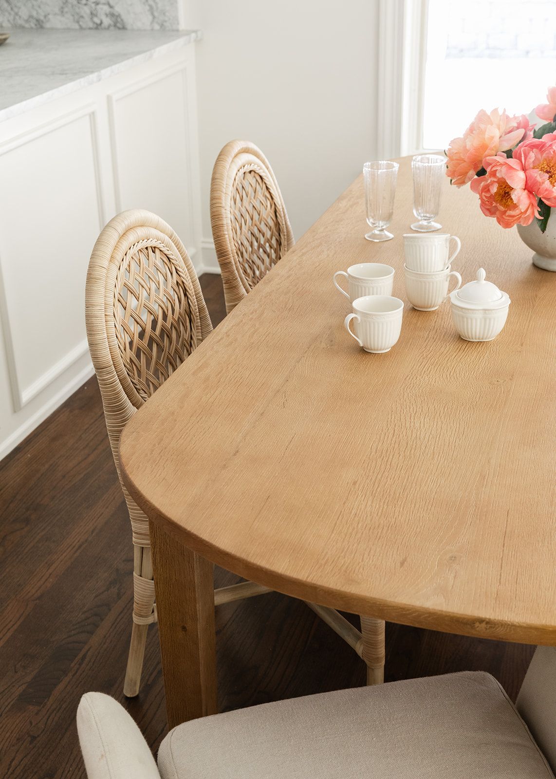 Oval oak dining table with rattan chairs, white tea set, and pink flowers in a bright dining room.