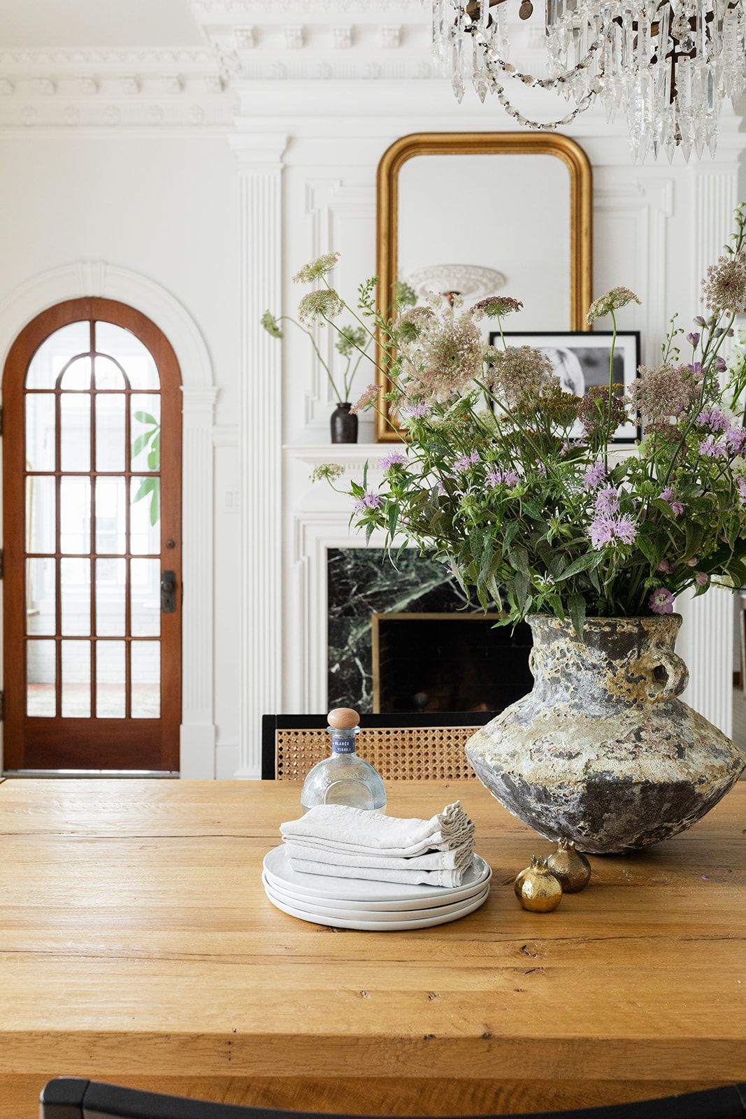 White dining room with marble fireplace, gold-framed mirror, and large floral arrangement on wooden table.