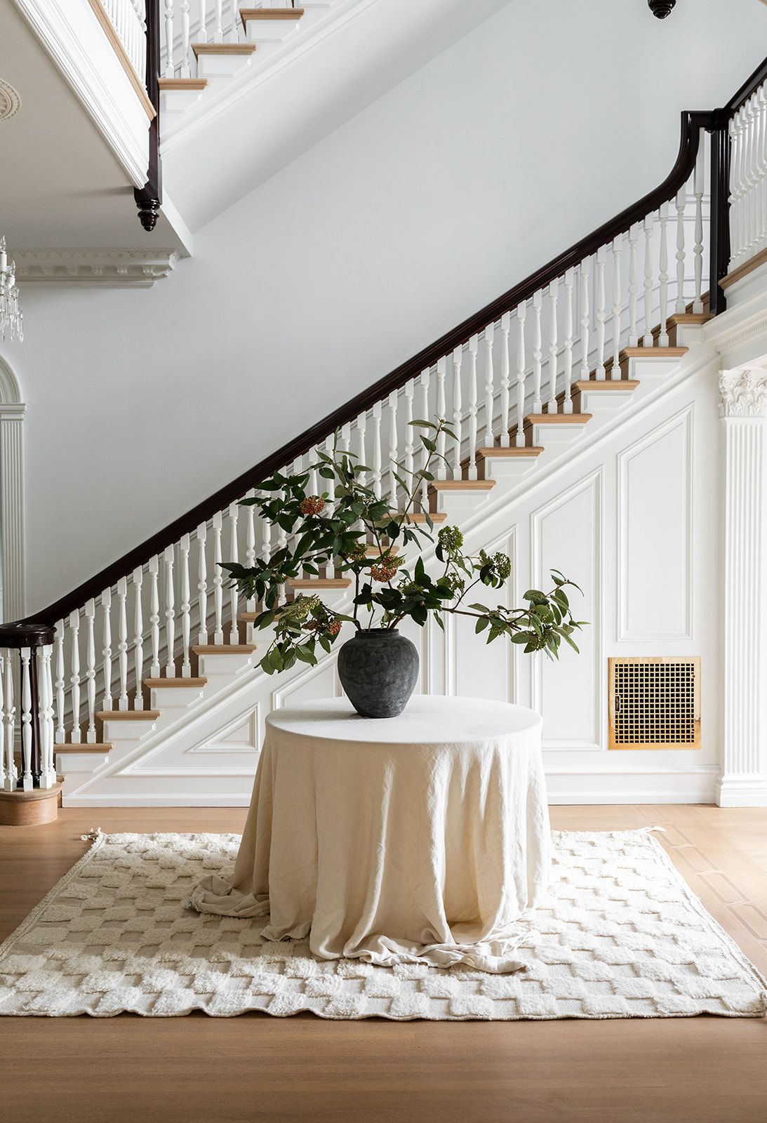 Grand white staircase with dark wood railings and a round table with floral arrangement on patterned rug.