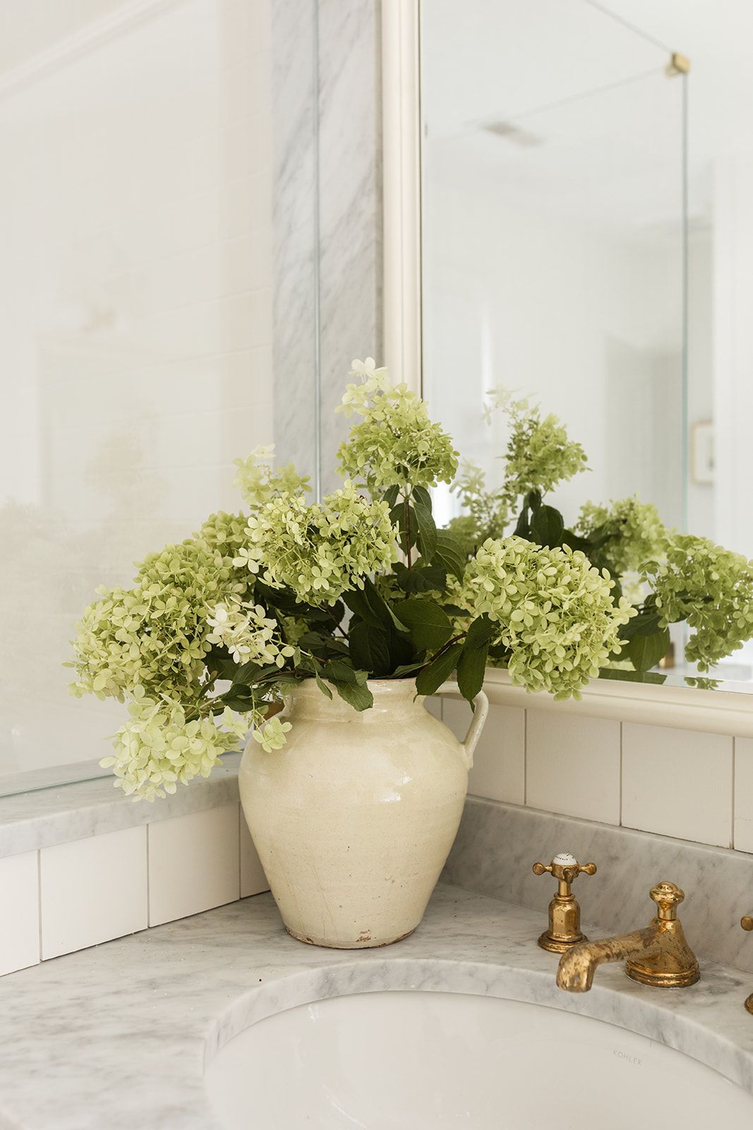 Large ceramic vase with fresh green hydrangeas on marble bathroom vanity beside antique brass faucet and mirror.