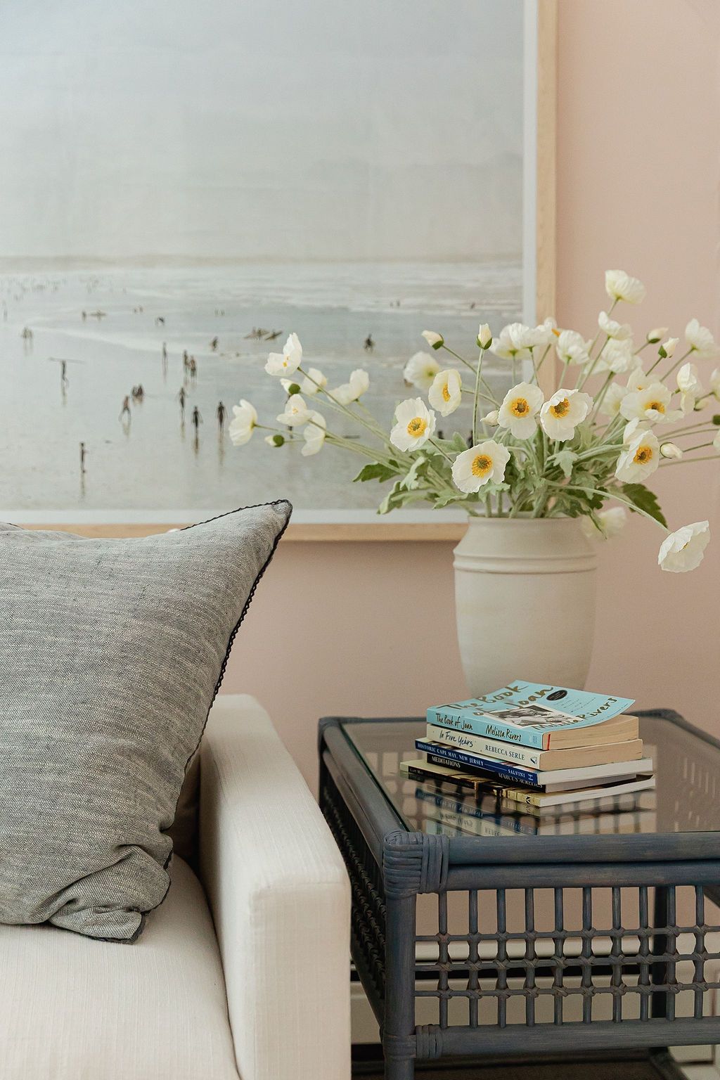 White flowers in a vase beside gray throw pillows and stacked books on a black metal table against a beige wall.