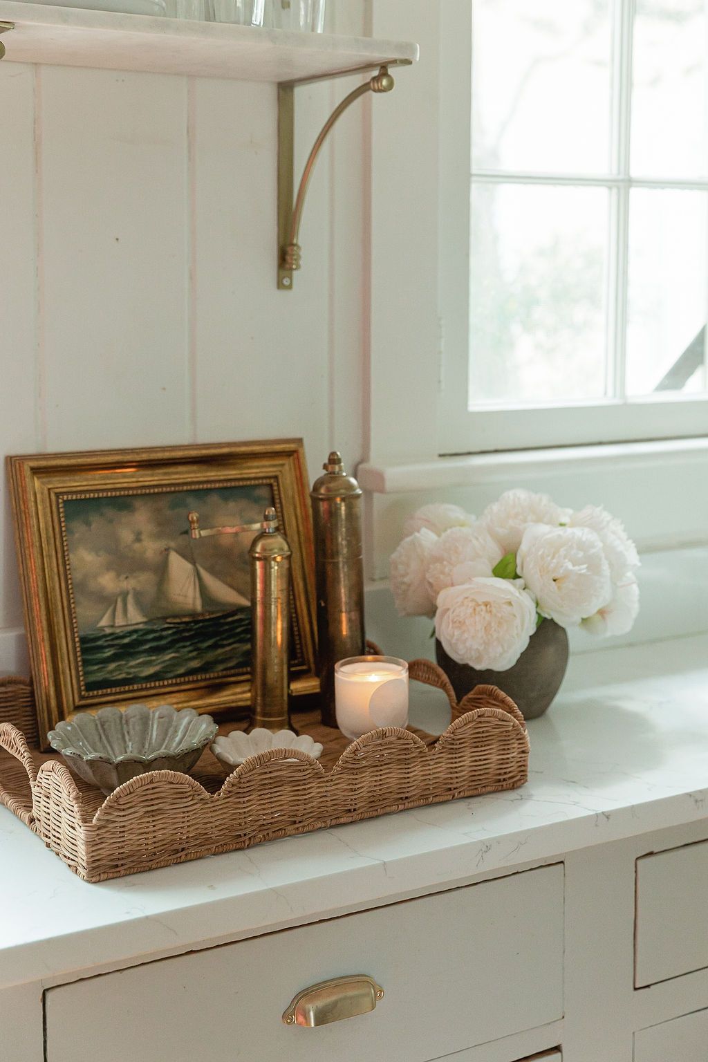 Coastal vignette with vintage sailing ship artwork, pink peonies, and wicker tray on white dresser by window.