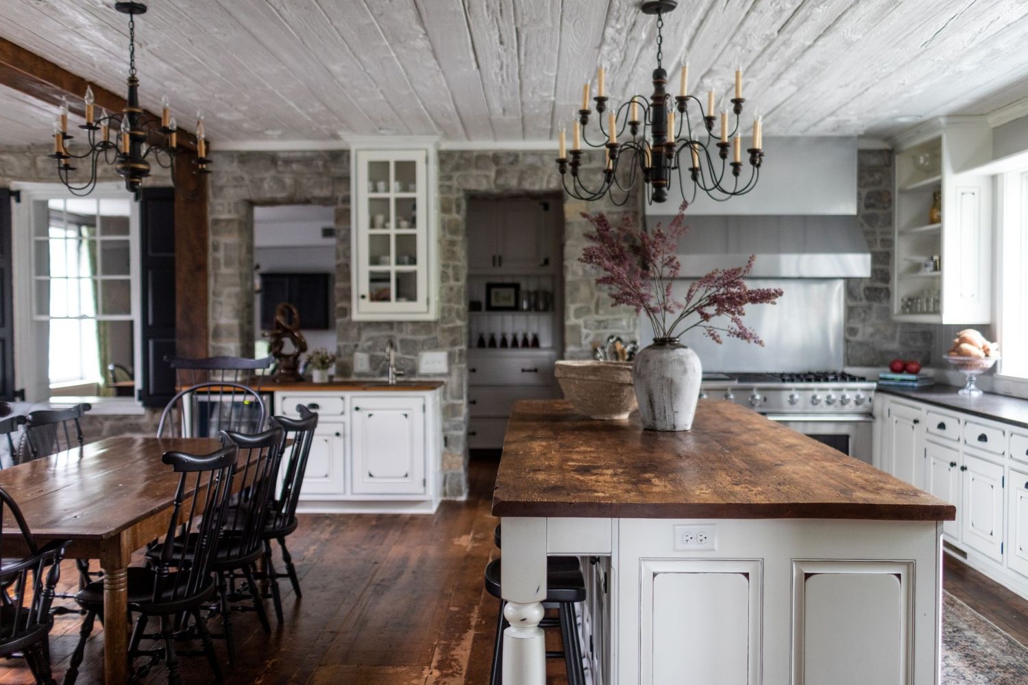 Rustic farmhouse kitchen with white cabinets, wooden countertops, ceiling chandeliers and dark wooden dining set.