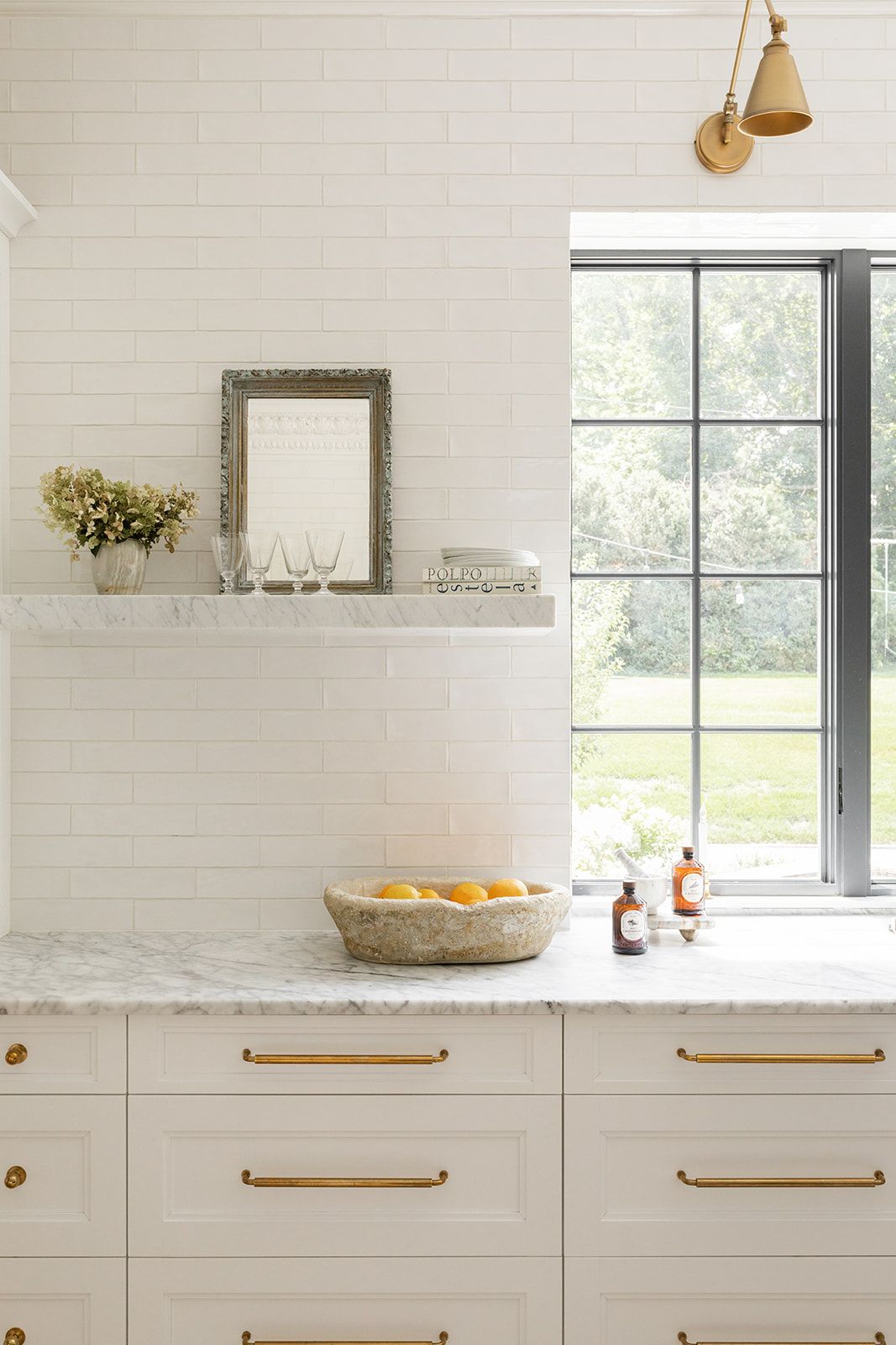 White kitchen with marble countertops, brass hardware, and large window overlooking green lawn.