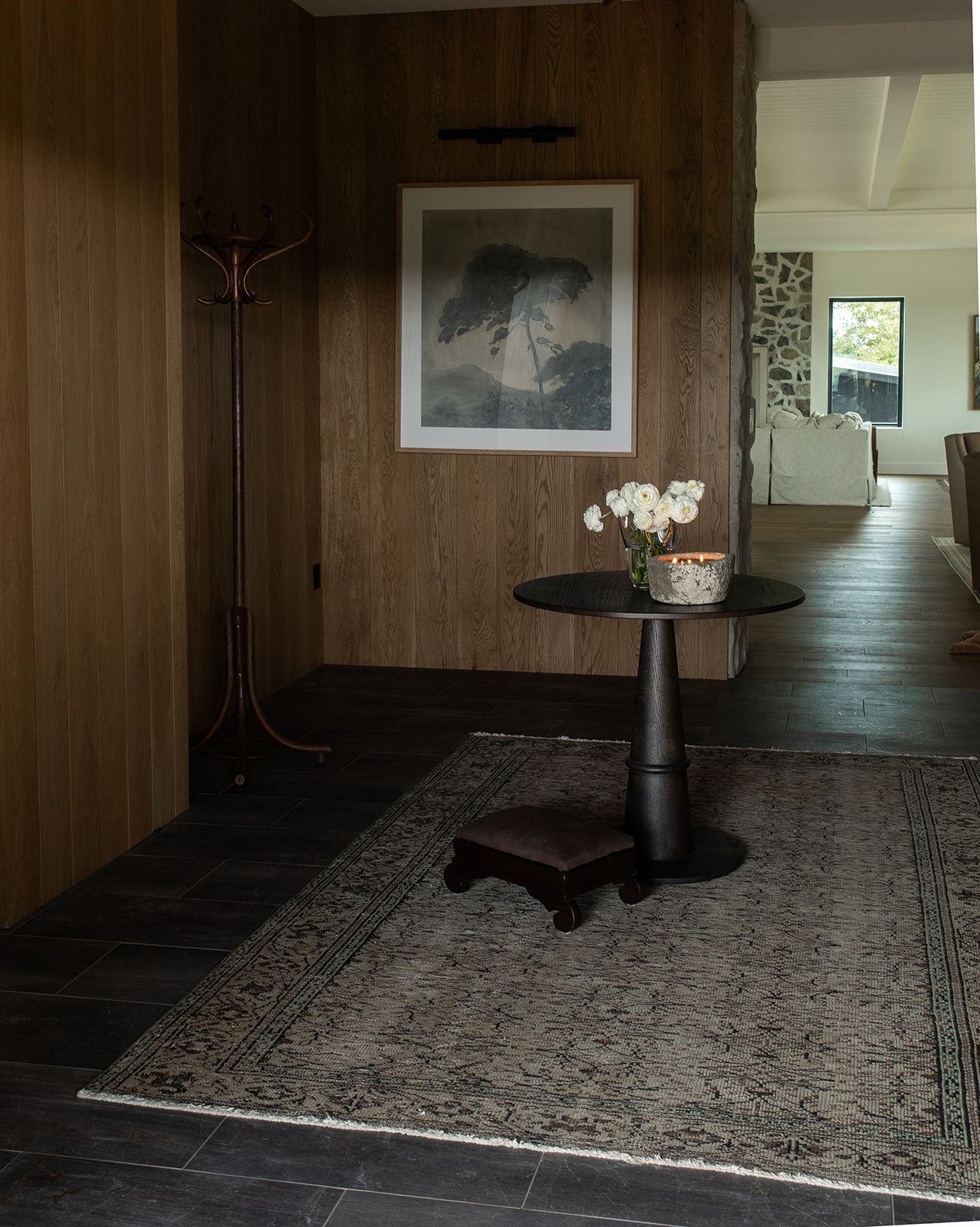 Elegant entryway with dark wood paneling, vintage rug, pedestal table with white flowers, and framed artwork.
