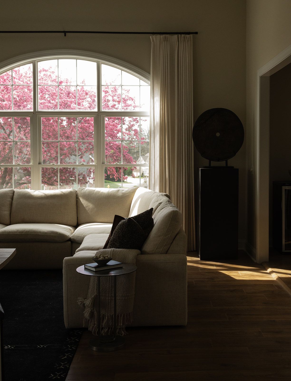Living room interior with arched windows framing pink blossoming trees and white upholstered furniture.
