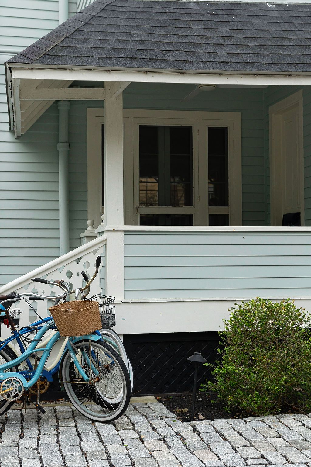 Blue bicycle parked on stone pavers beneath a white porch overhang of a light blue house.