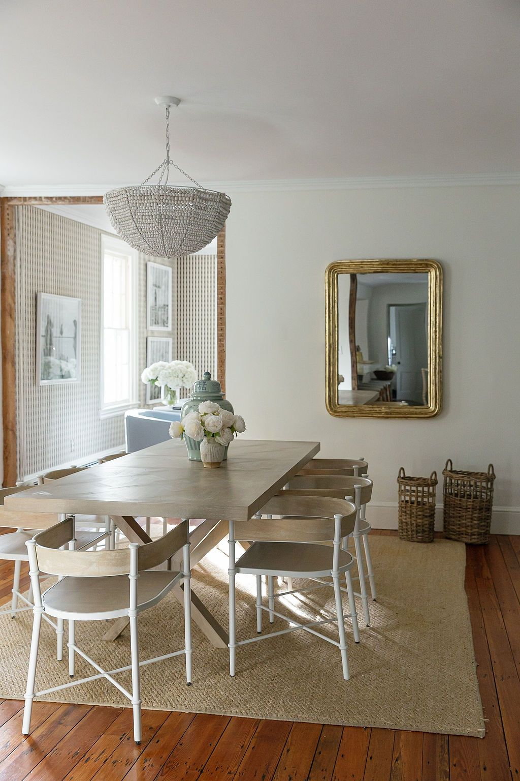 Coastal dining room with beaded chandelier, rustic wood table, white chairs, gold mirror and natural jute rug.