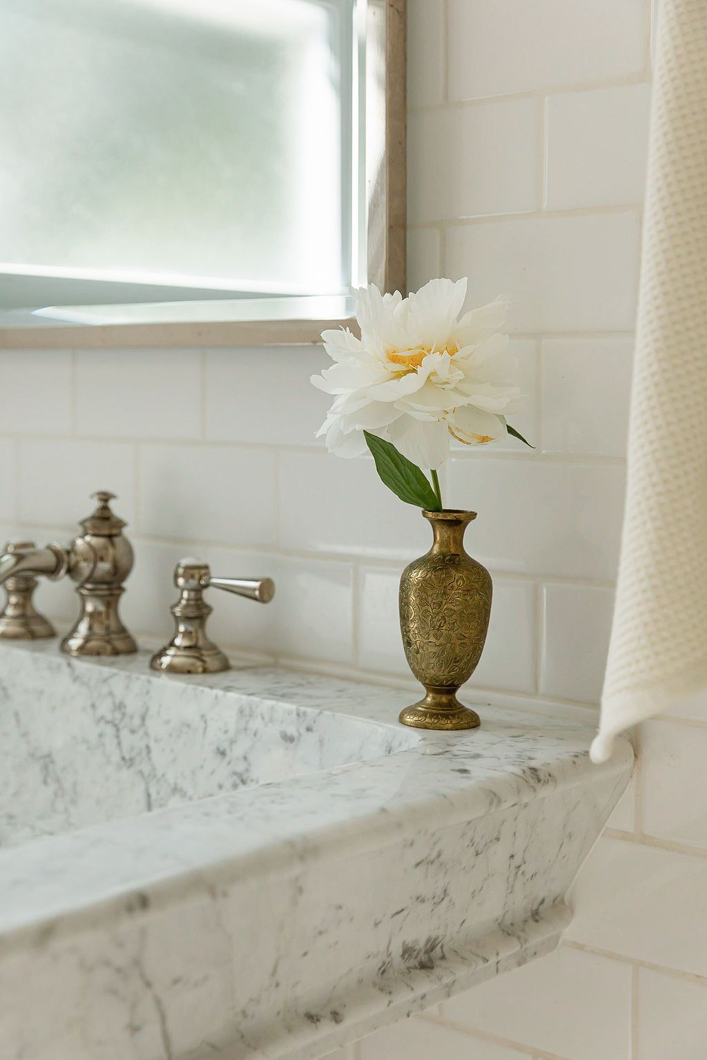 A delicate white flower sits in a brass vase on a marble bathroom counter near vintage-style faucets and window.
