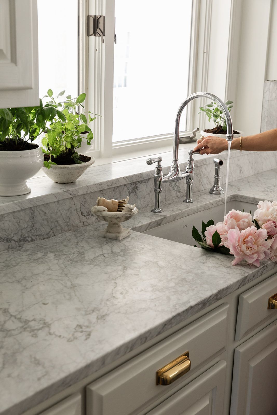 Elegant marble kitchen countertop with chrome bridge faucet, fresh herbs, and pink peony flowers near sink.