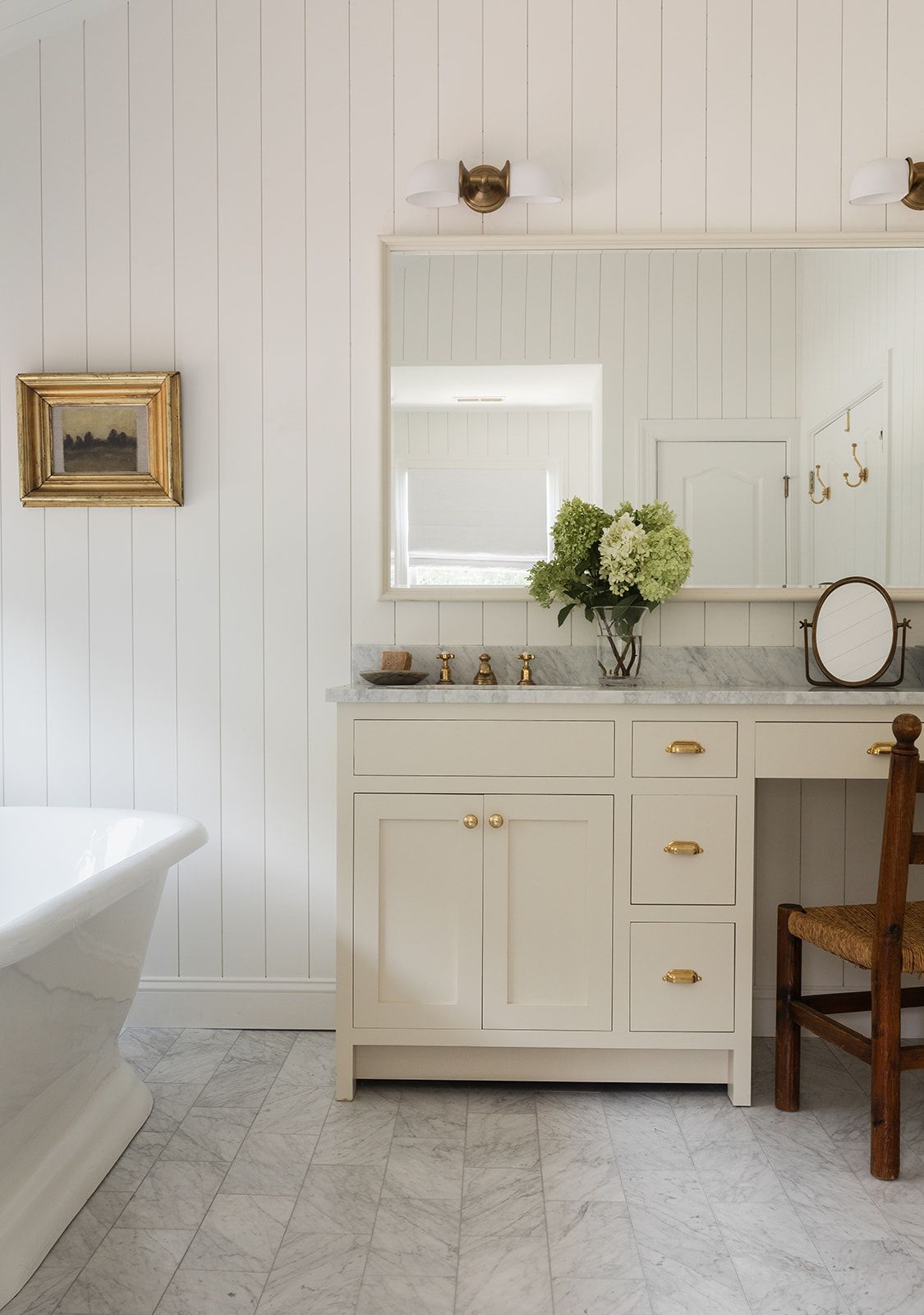 White beadboard bathroom with cream vanity, brass fixtures, marble countertop, and vintage gold-framed artwork above a freestanding tub.