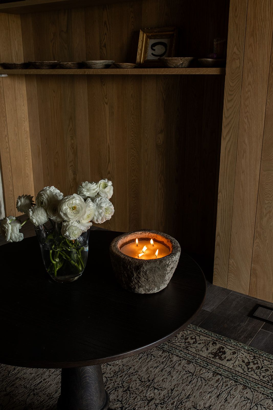 A lit multi-wick candle in a stone vessel beside white roses on a dark round table near wooden shelving.