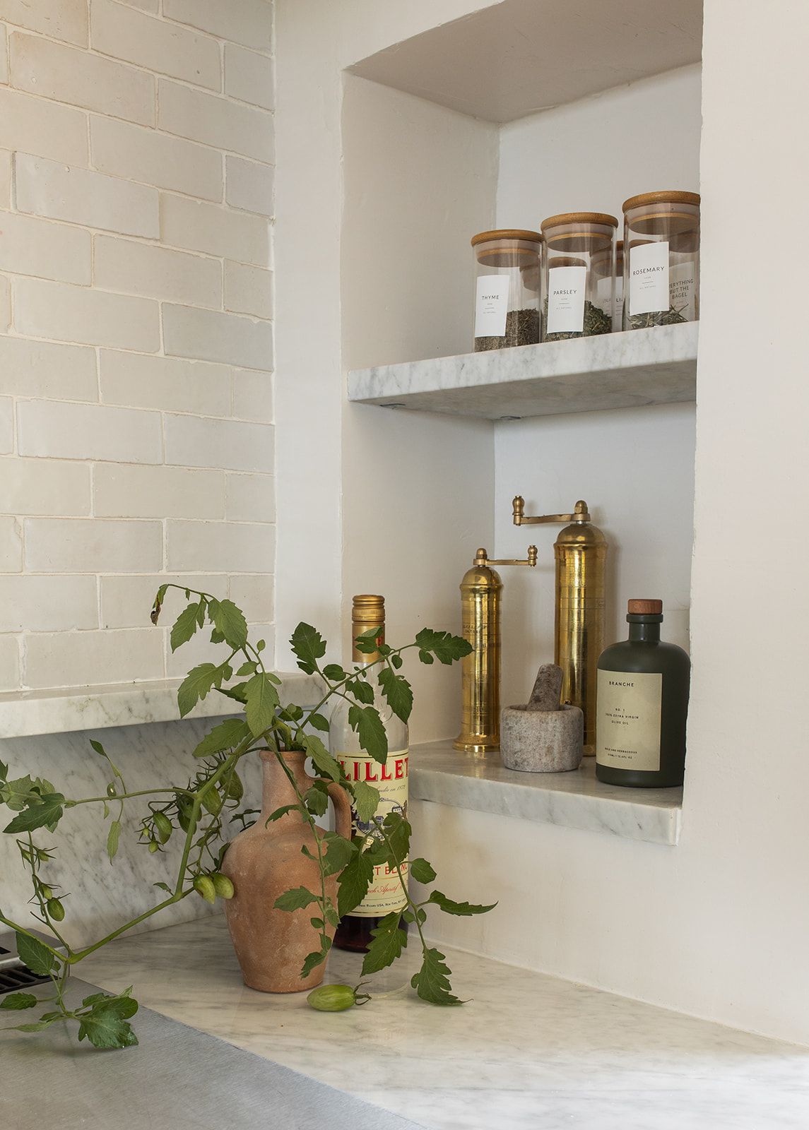 Marble kitchen niche shelves with glass spice jars, brass pepper mill, and olive oil bottle beside a terracotta plant pot.
