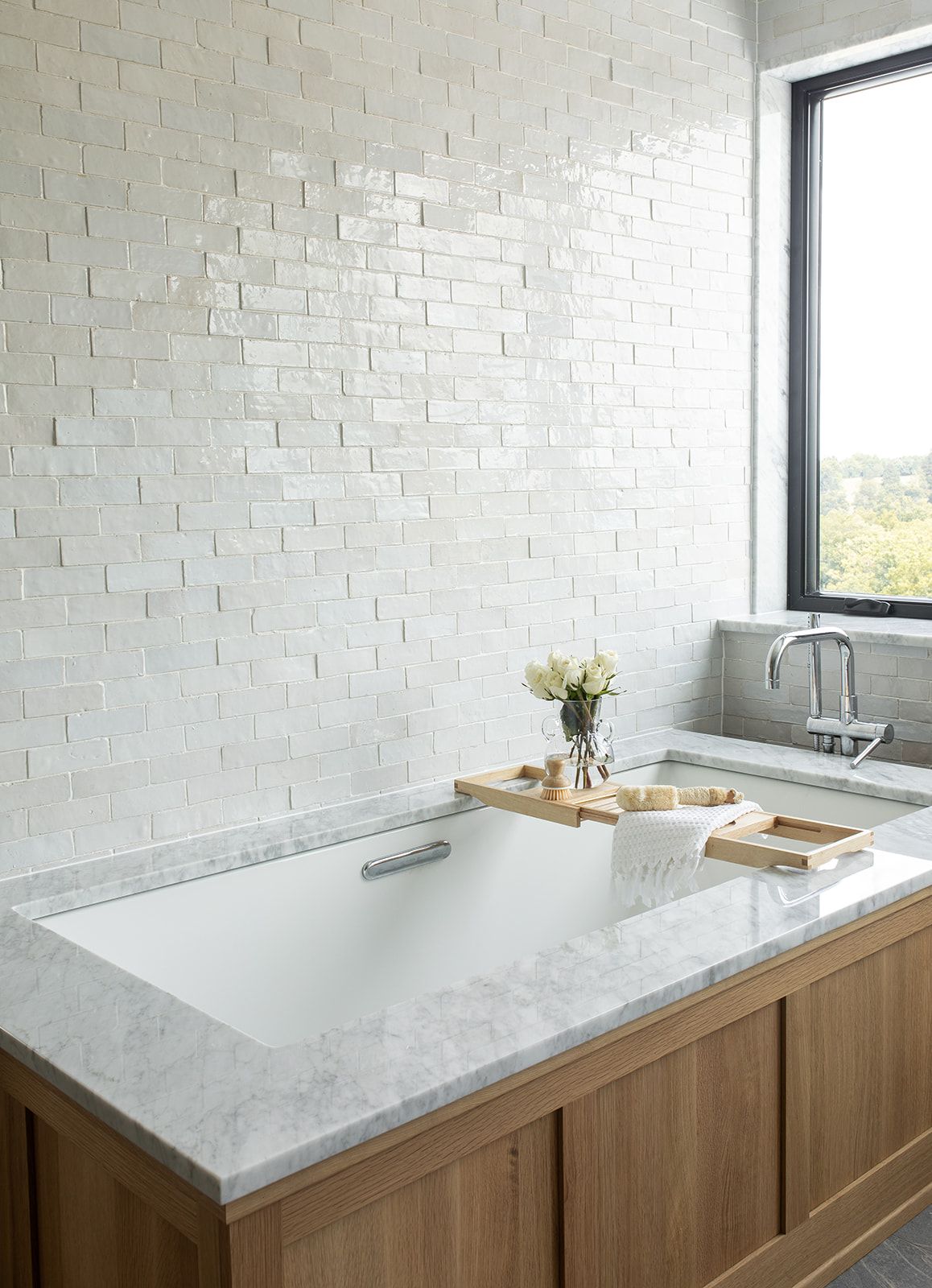 Luxury bathroom with white brick tile walls, wood vanity, marble countertop bathtub, and bamboo bath caddy.