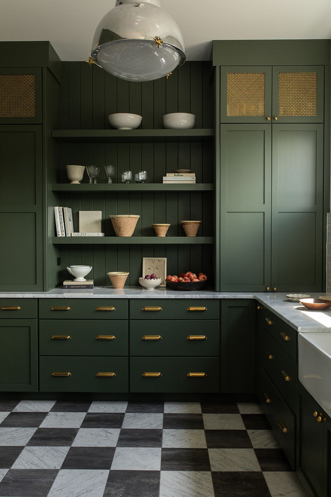 Dark green kitchen with brass hardware, checkered floor, open shelving, and rattan cabinet doors under a dome pendant light.