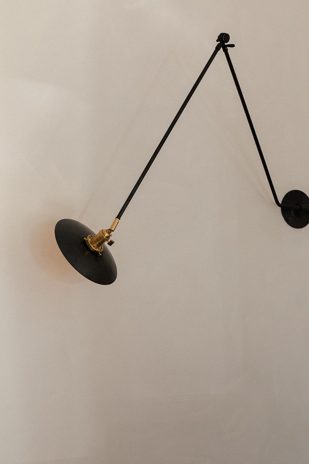 A minimalist black adjustable wall lamp with brass details extends from a beige wall on an articulating arm.