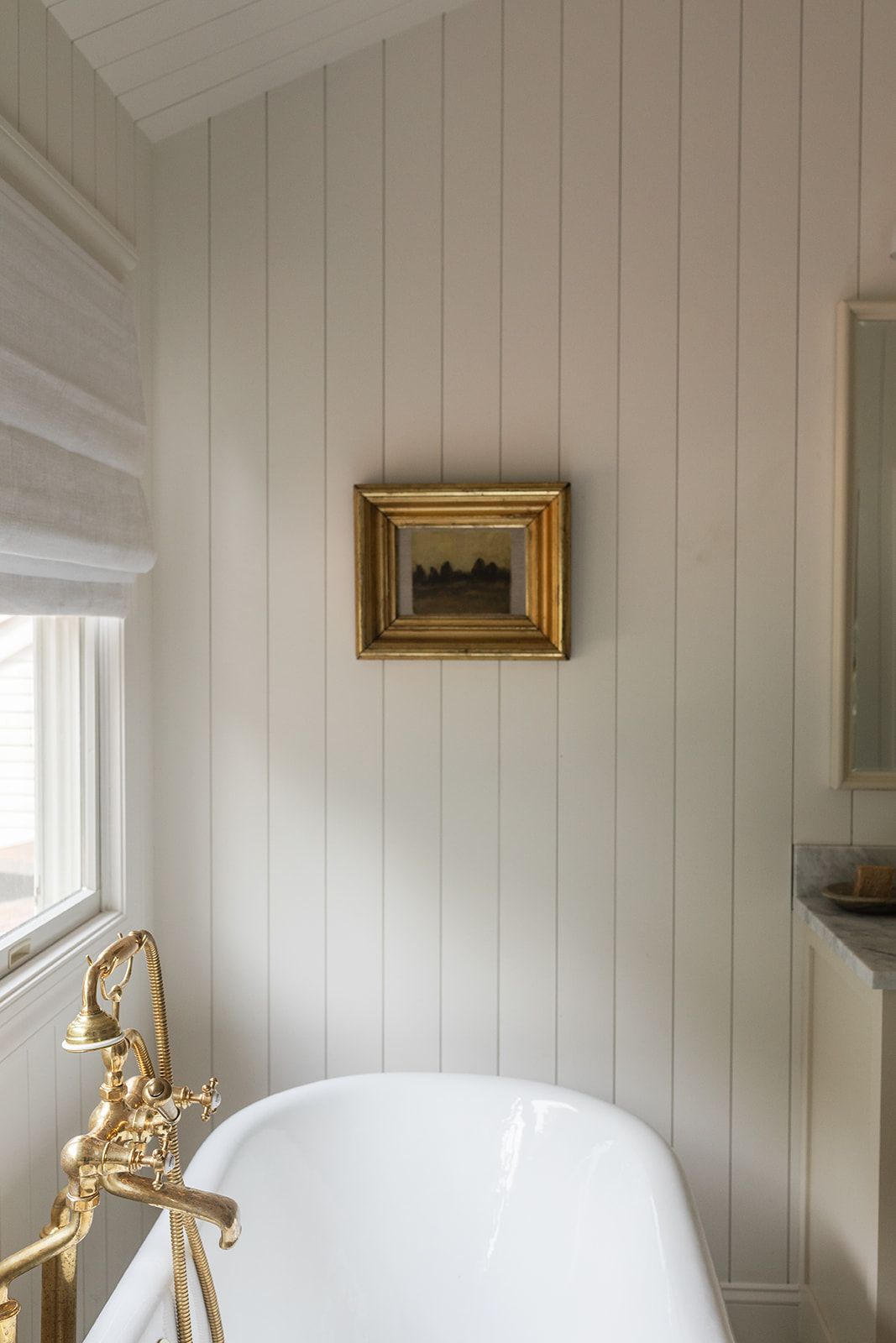 Serene white beadboard bathroom with freestanding tub, ornate brass floor faucet, and vintage gold-framed landscape painting.