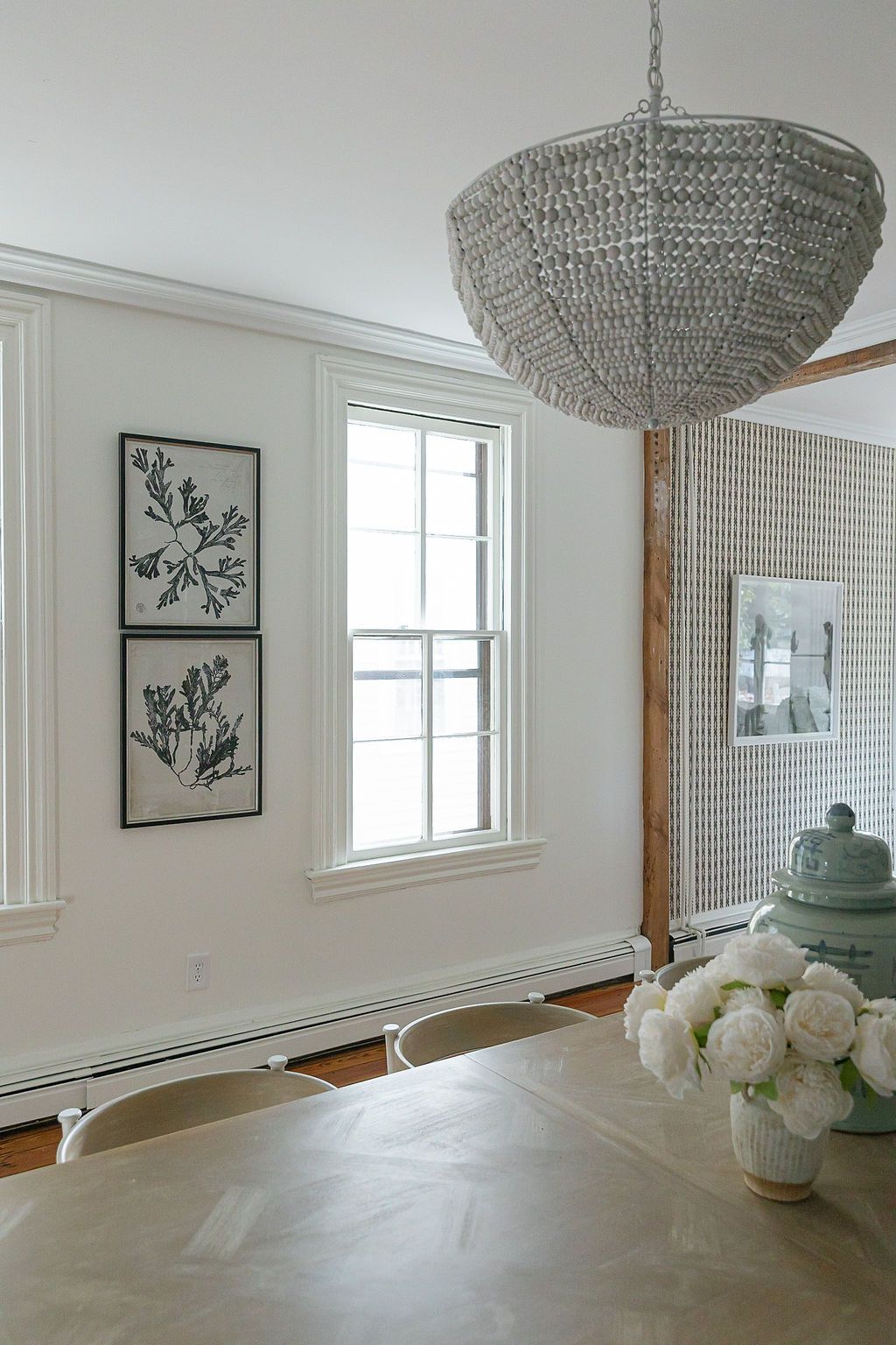 Elegant dining room with crystal chandelier, framed botanical prints, and white roses in a modern coastal-inspired setting.