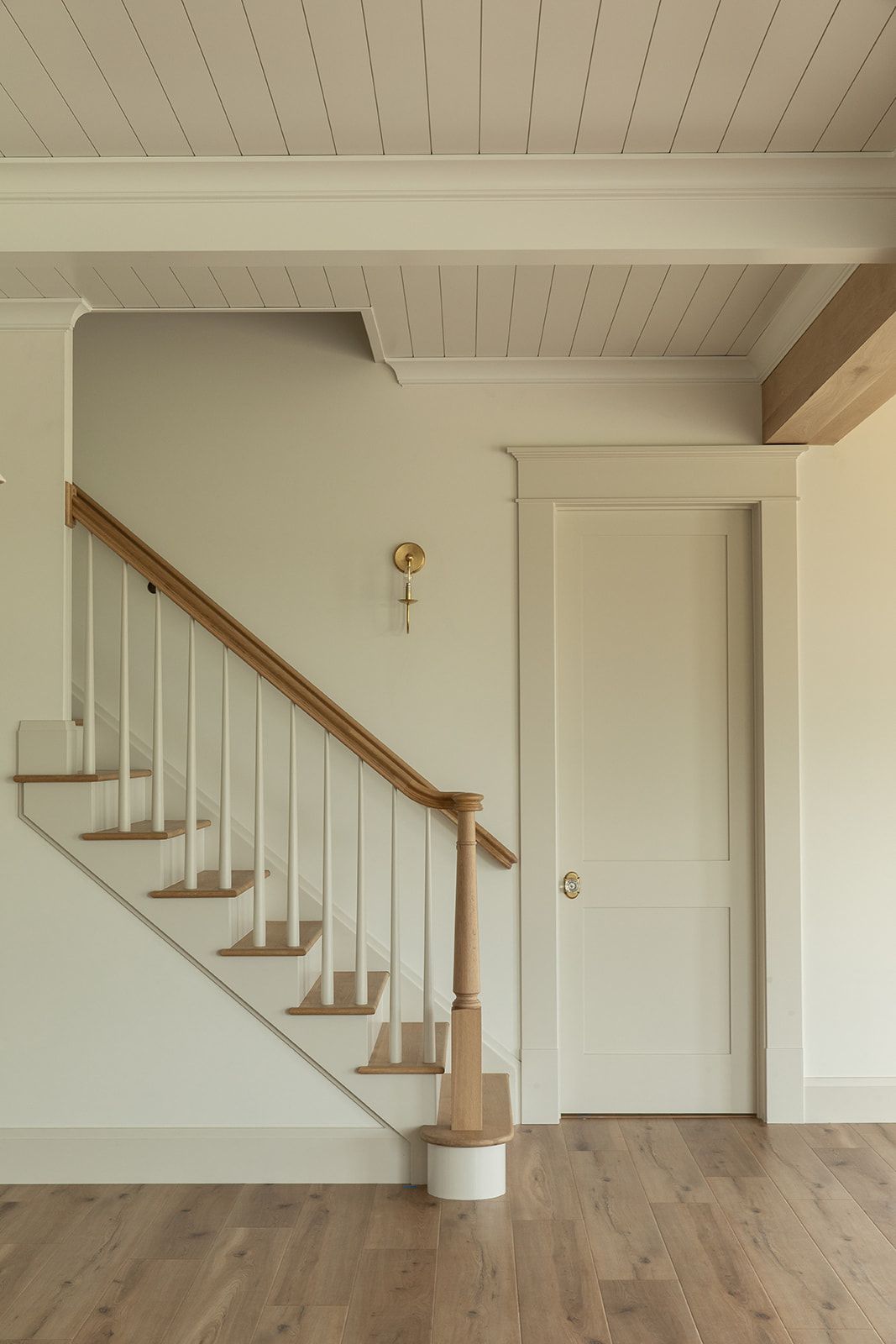 Elegant foyer with white staircase, wood handrail, shiplap ceiling, brass wall sconce, and light hardwood floors.