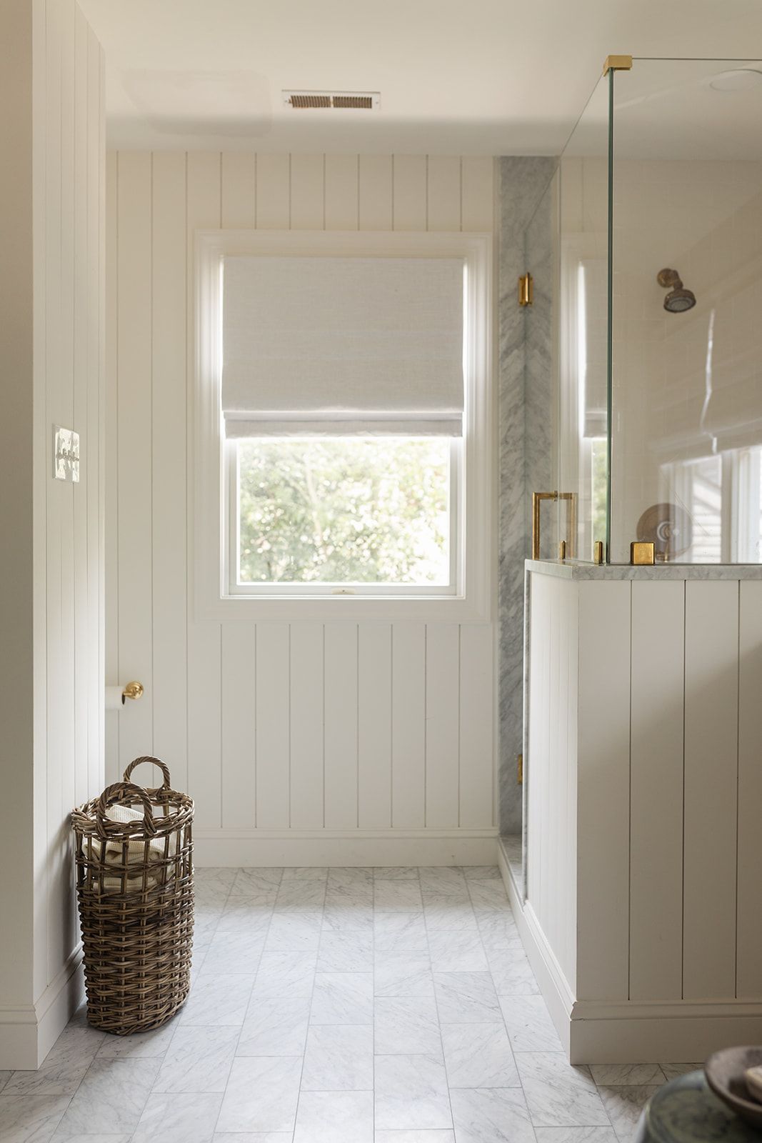 Bright white beadboard bathroom with marble tile floor, glass shower, brass hardware, and woven basket near window.