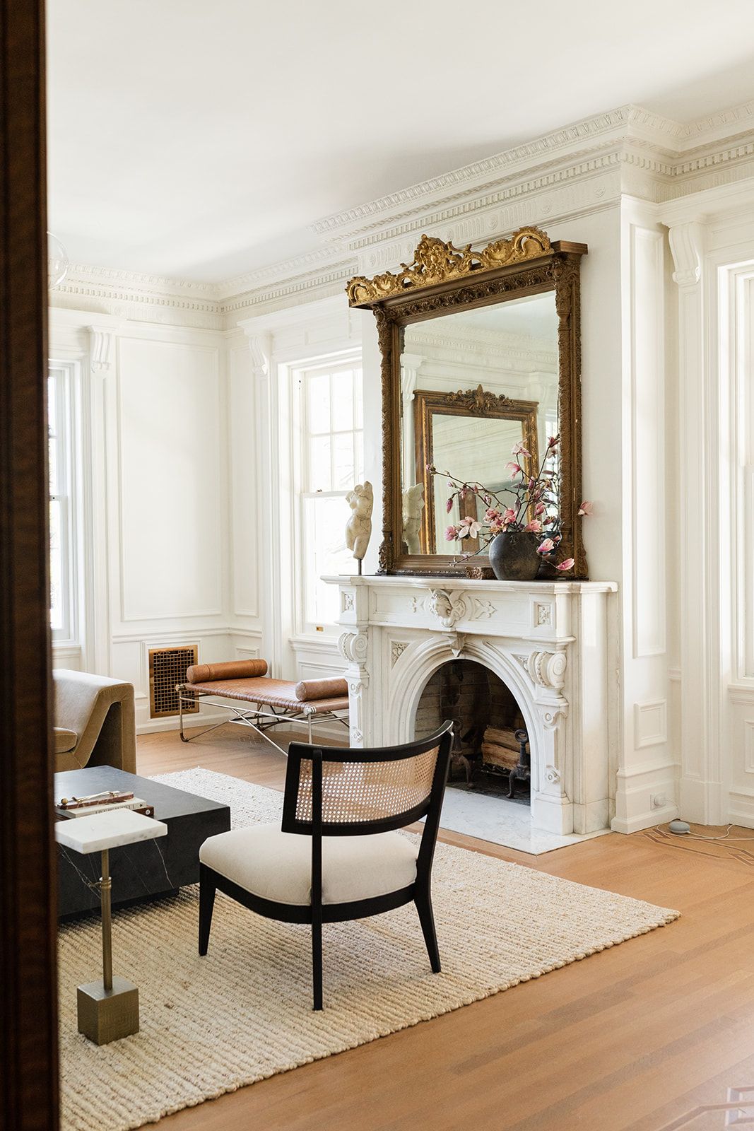 Elegant living room with ornate gold mirror above white marble fireplace and modern black accent chair.
