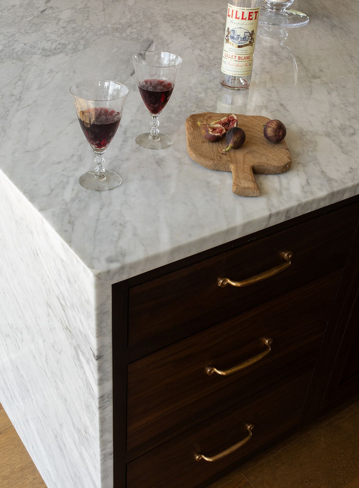 Marble kitchen island with dark wood drawers, brass handles, two red wine glasses, cutting board, and a bottle of Lillet.