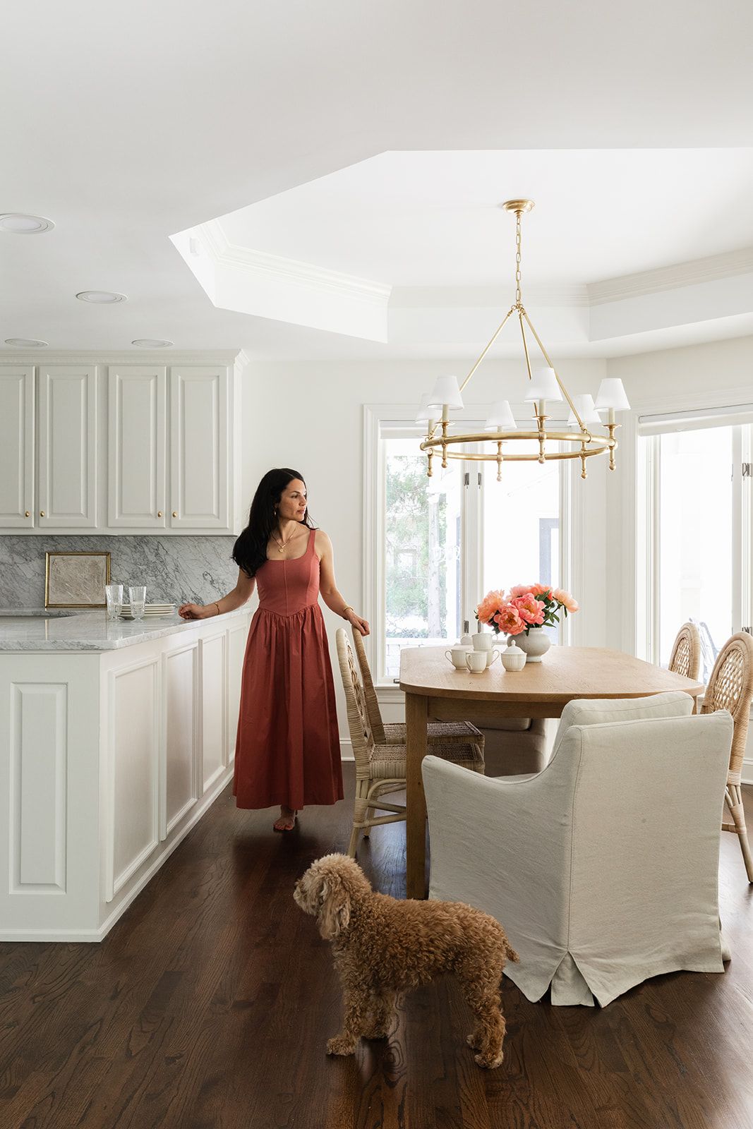 Woman in red dress stands in white kitchen beside oval wood dining table with gold chandelier and a brown doodle dog.