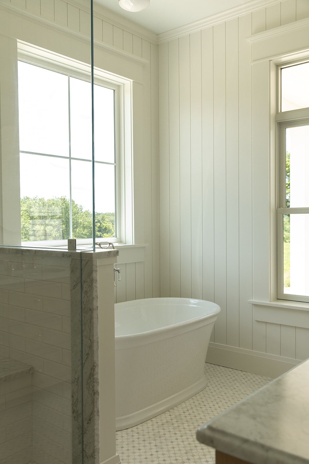 White freestanding soaking tub beside shiplap walls and large windows in a bright, airy bathroom with mosaic tile floors.