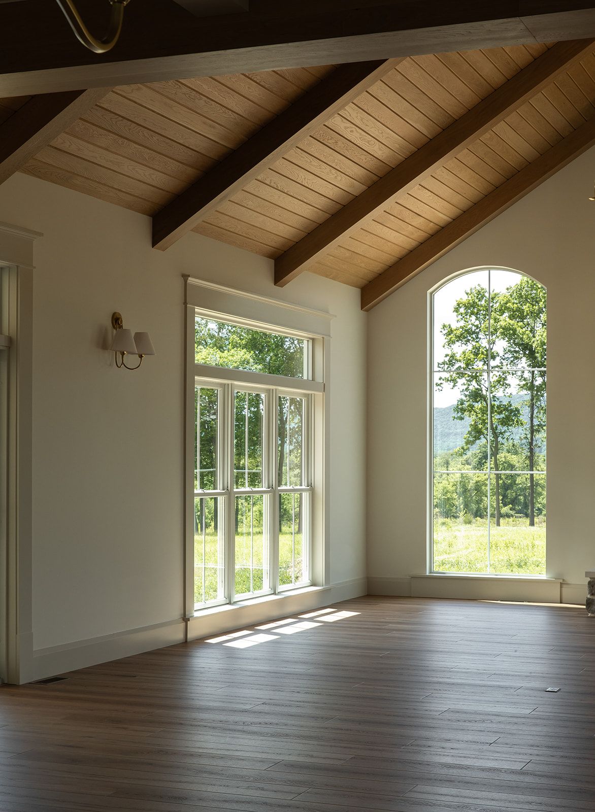 Empty room with vaulted wood-beam ceiling, hardwood floors, and large arched windows overlooking lush green landscape.